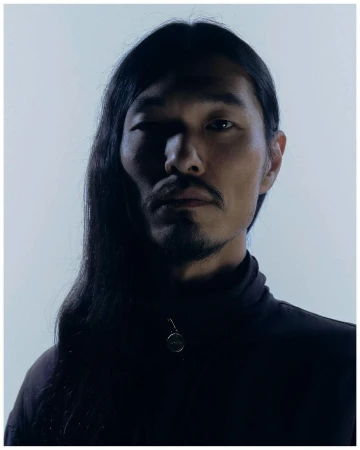 Portrait of a man with long dark hair and facial hair wearing a dark zip-up jacket against a plain light background