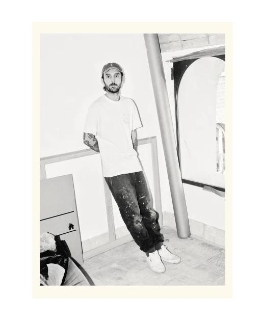 Monochrome portrait of a man in casual clothing leaning against a wall next to framed artwork, wearing a cap and jeans with paint splatters.