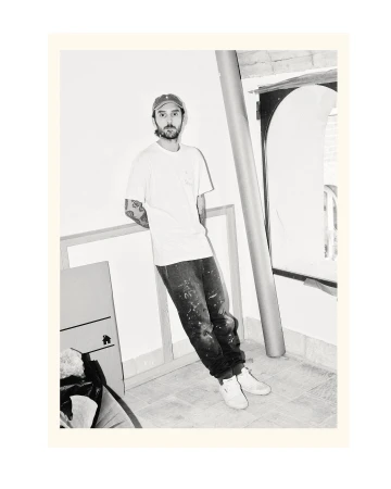 Monochrome portrait of a man in casual clothing leaning against a wall next to framed artwork, wearing a cap and jeans with paint splatters.