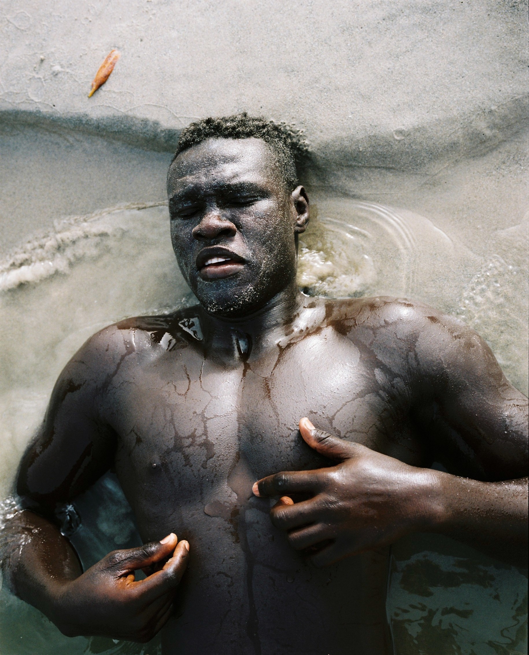 Adult male lies in shallow water with hands near his chest. The dark-skinned man has his eyes closed, with a leaf visible near his head.