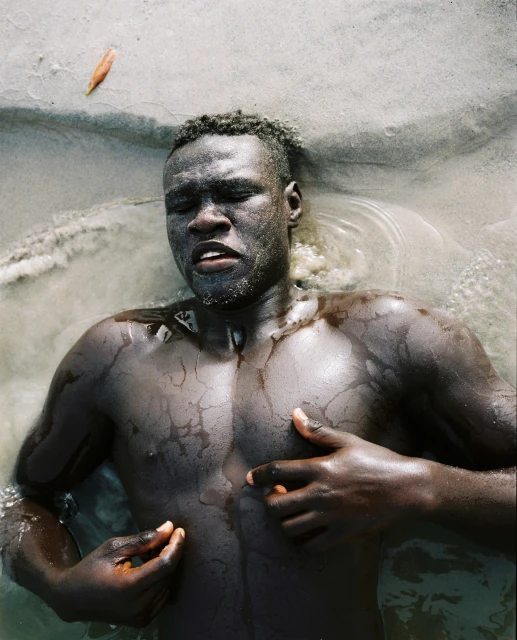 Adult male lies in shallow water with hands near his chest. The dark-skinned man has his eyes closed, with a leaf visible near his head.