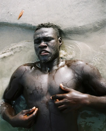 Adult male lies in shallow water with hands near his chest. The dark-skinned man has his eyes closed, with a leaf visible near his head.
