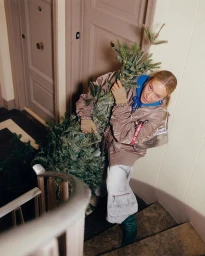 Woman in bomber jacket and light grey jeans holding a small pine tree while standing on a staircase, photographed by Dani Pujalte.