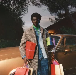 Black man holding colourful shopping boxes and bags, wearing a houndstooth coat, denim jacket and green turtleneck.