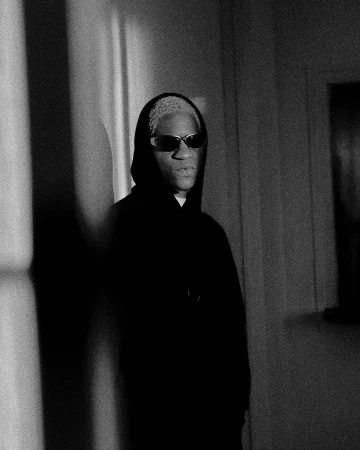 Monochrome portrait of a Black man with short light hair wearing dark sunglasses and a black hoodie in a sunlit interior.