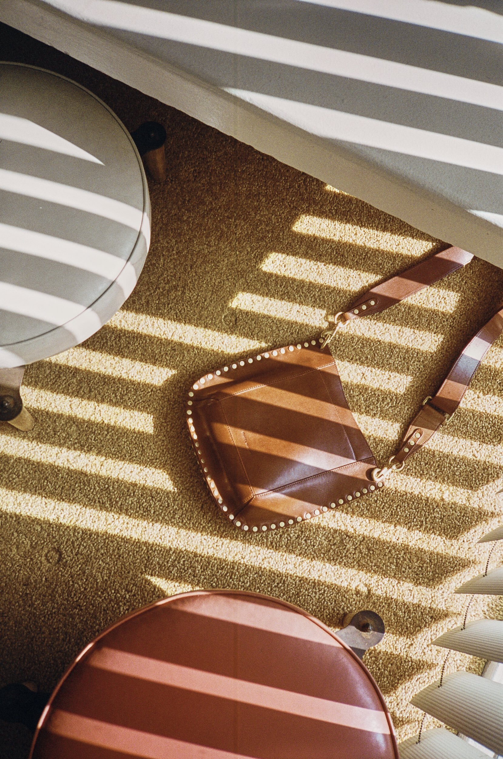 Overhead shot of a brown striped purse on a carpet floor, flanked by two round stools and shadowed by window blinds.