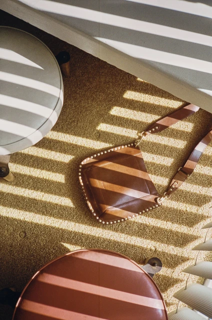 Overhead shot of a brown striped purse on a carpet floor, flanked by two round stools and shadowed by window blinds.