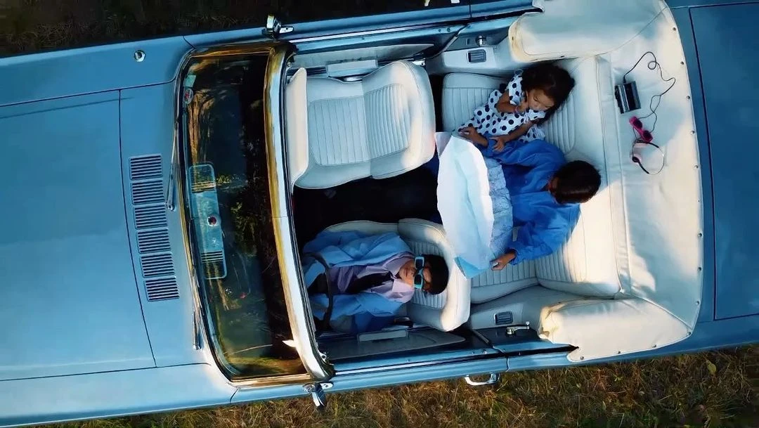 Overhead shot of two children relaxing in a light blue convertible car on a grassy field, capturing a carefree moment.