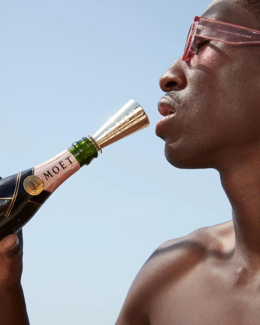 Adult male in sunglasses drinks from a champagne bottle outdoors against a clear blue sky in a medium shot.