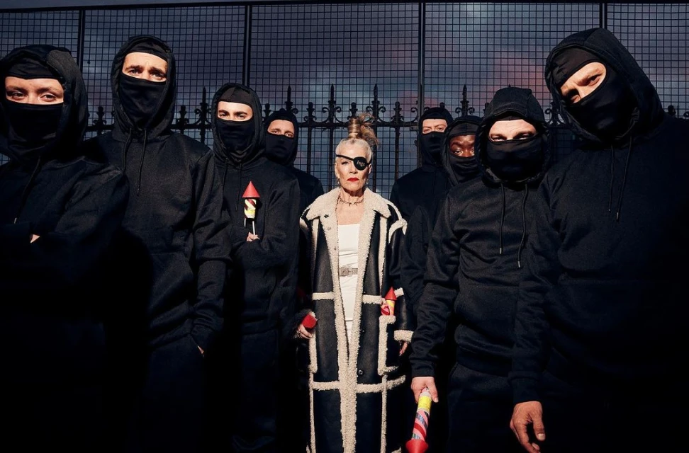 Editorial shot of an adult woman with an eye patch standing in front of a group of people wearing black ski masks against a fence.
