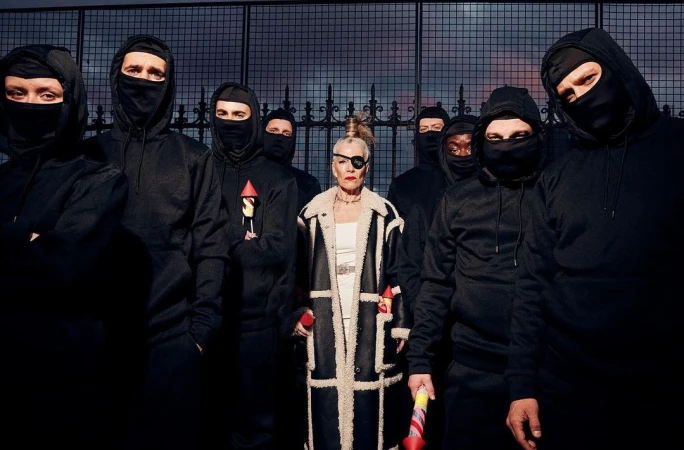 Editorial shot of an adult woman with an eye patch standing in front of a group of people wearing black ski masks against a fence.