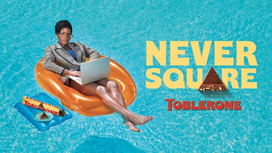 Woman in blazer works on a laptop while floating on an orange inflatable in a pool, beside Toblerone chocolate bar and promotional text.