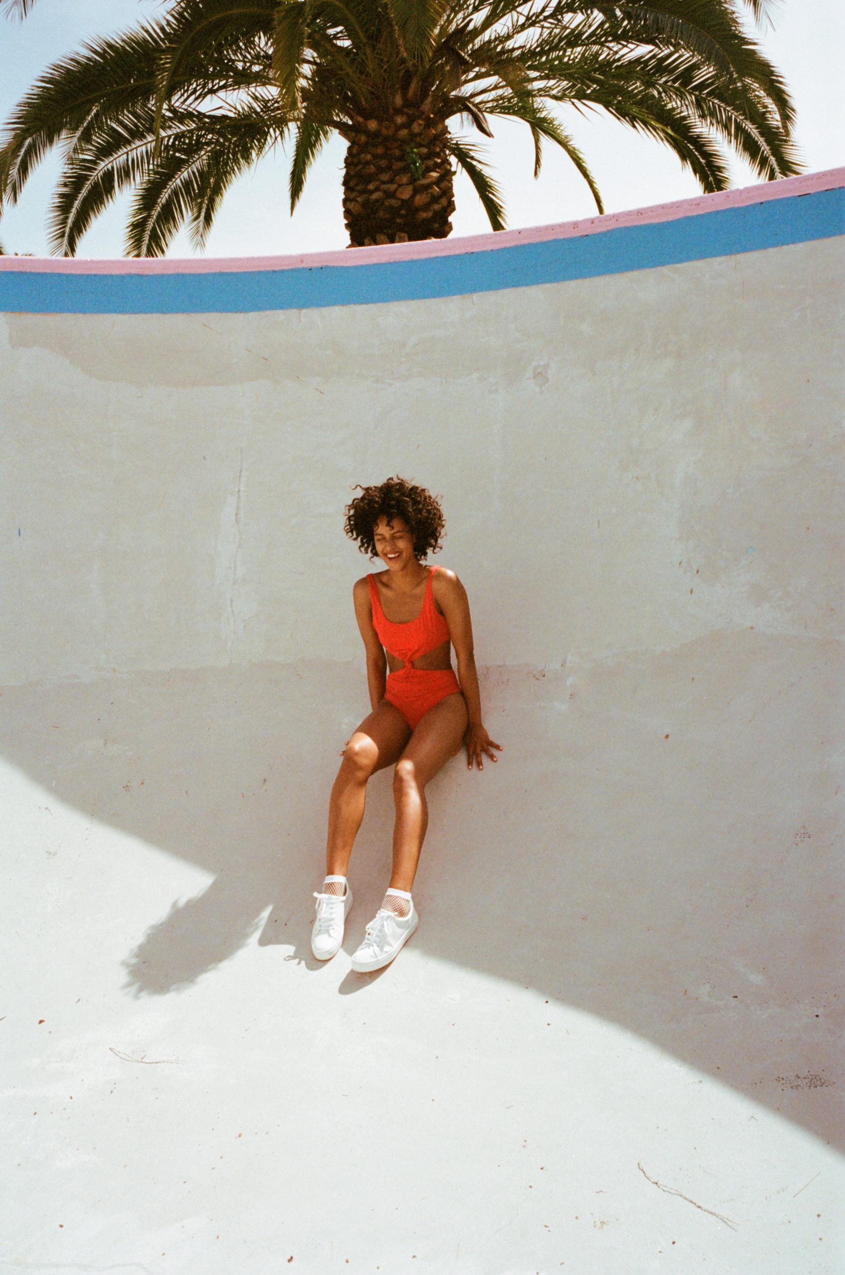 Woman in red one-piece swimsuit and white sneakers seated inside an empty concrete skate bowl beneath a palm tree.