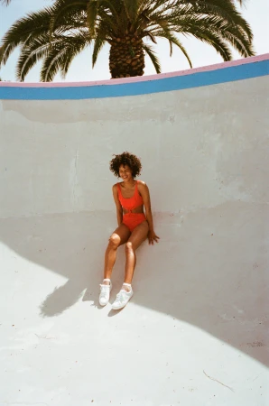 Woman in red one-piece swimsuit and white sneakers seated inside an empty concrete skate bowl beneath a palm tree.