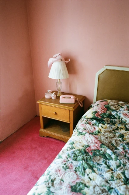 Bedroom interior with a floral bedspread, pink walls and carpet, a wooden nightstand with a lamp, telephone, and a pink cowboy hat.
