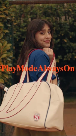 Brunette woman with a tennis bag slung over her shoulder, in an outdoor park-like setting with greenery, wearing a blue t-shirt.