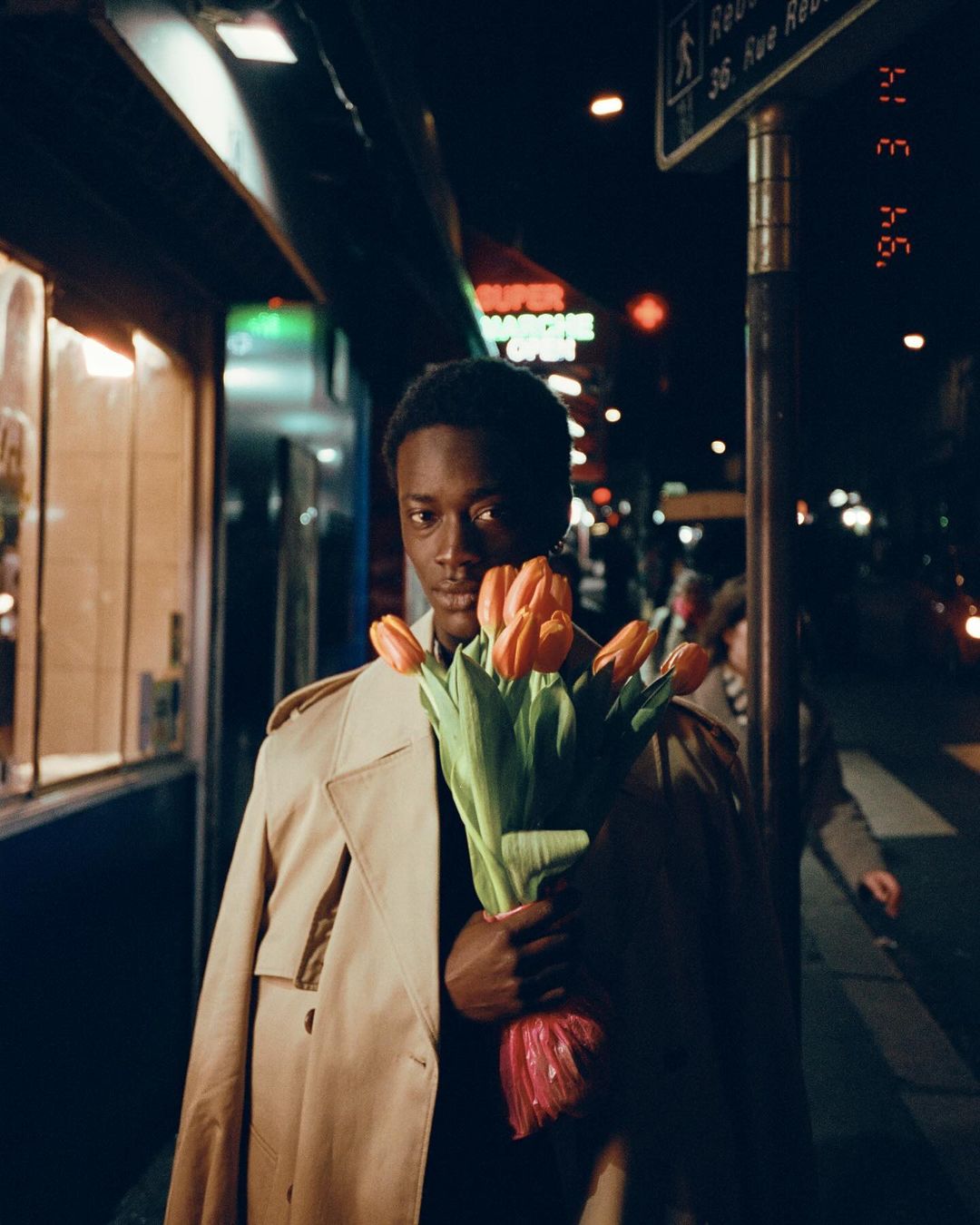 Stylish man in trench coat holds orange tulips on a city street at night, illuminated by storefront and street lights, creating a moody ambiance.