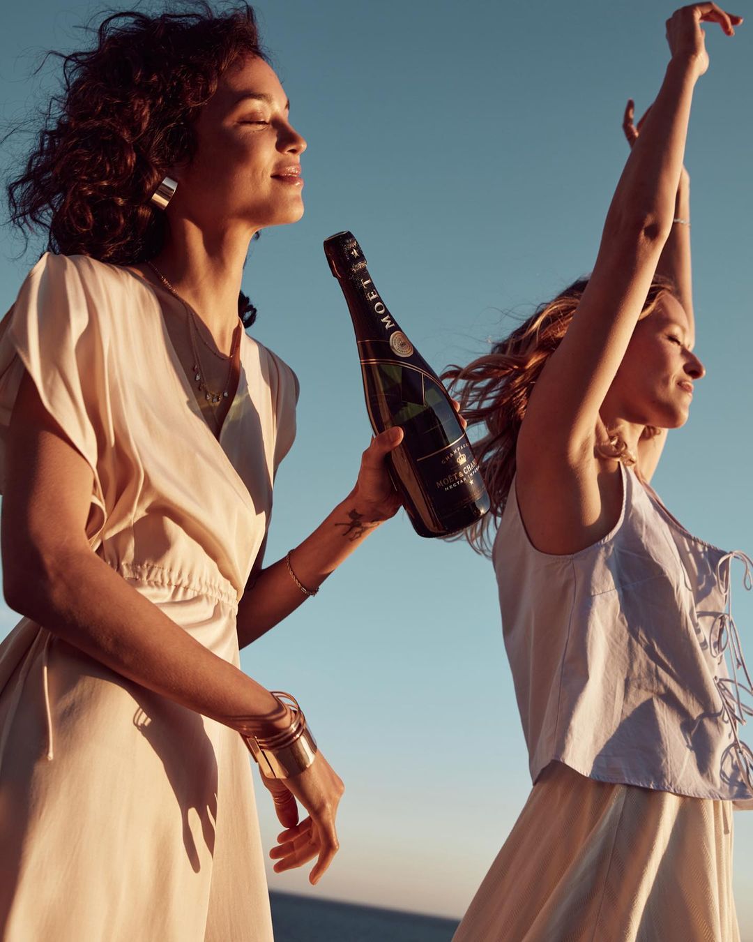 Two women celebrate with champagne on a beach, bathed in warm sunlight, creating a celebratory and carefree atmosphere with a clear sky backdrop.