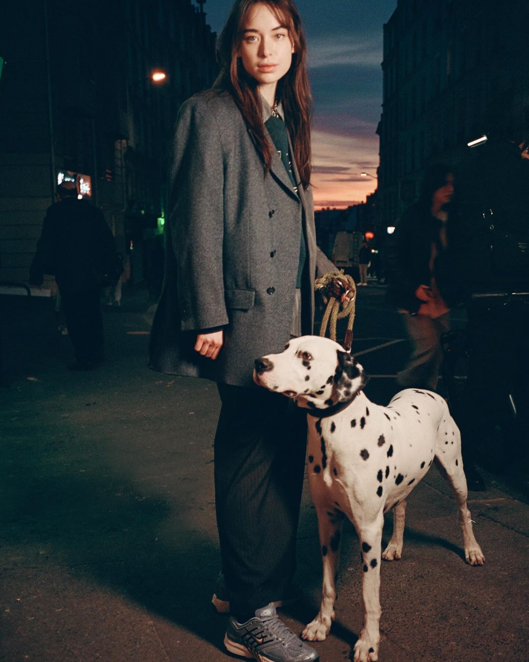 A woman and a dalmatian dog stand on an urban street at dusk. She is wearing a grey coat and sneakers. City buildings in the background.