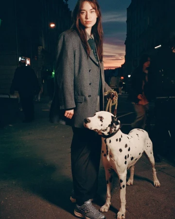 A woman and a dalmatian dog stand on an urban street at dusk. She is wearing a grey coat and sneakers. City buildings in the background.