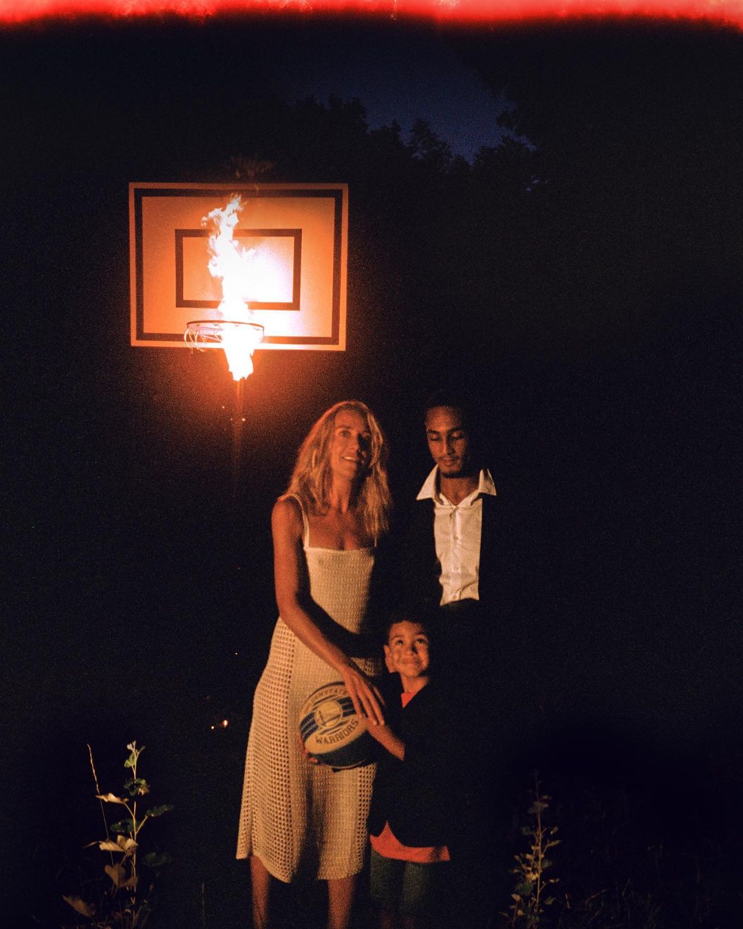 A family of three stands in a dark outdoor setting in front of a basketball hoop engulfed in flames, creating a striking and somewhat surreal portrait.