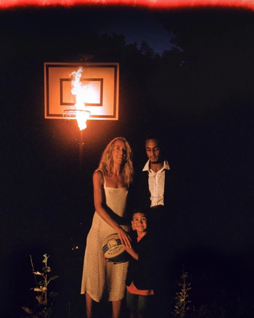 A family of three stands in a dark outdoor setting in front of a basketball hoop engulfed in flames, creating a striking and somewhat surreal portrait.