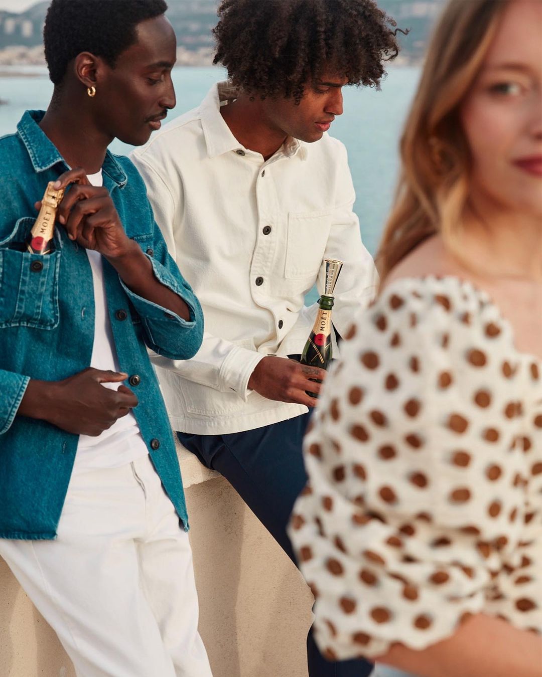 Three people in casual clothing are together outside with bottles of champagne, with a blurred out woman to the right.