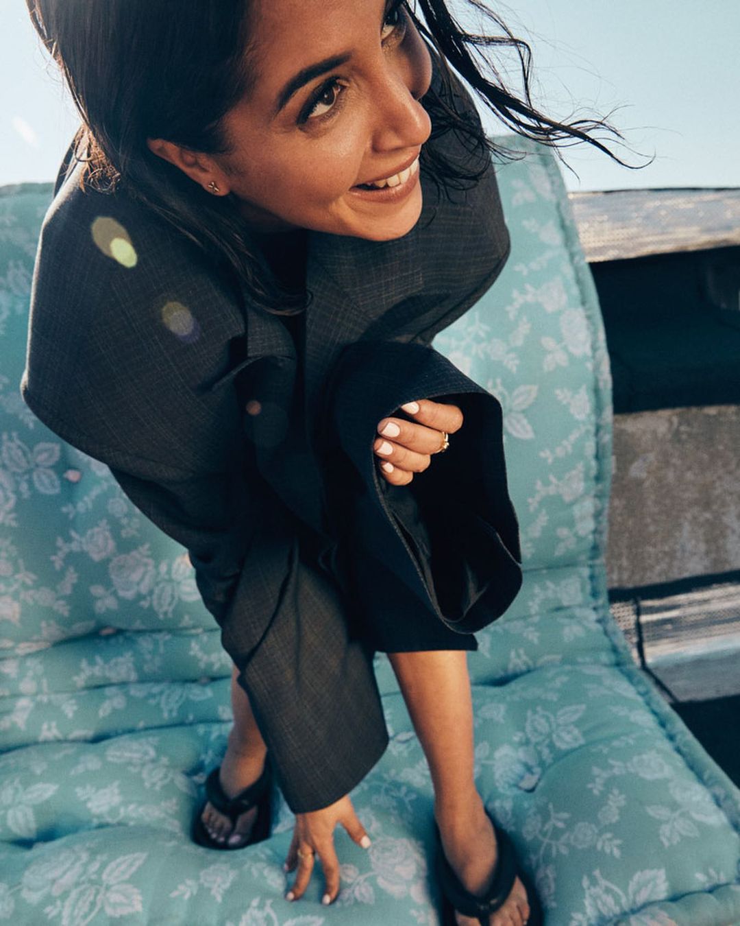 Attractive woman in suit poses on floral cushion outdoors with bright sunlight, showcasing a candid, fashion-forward aesthetic.