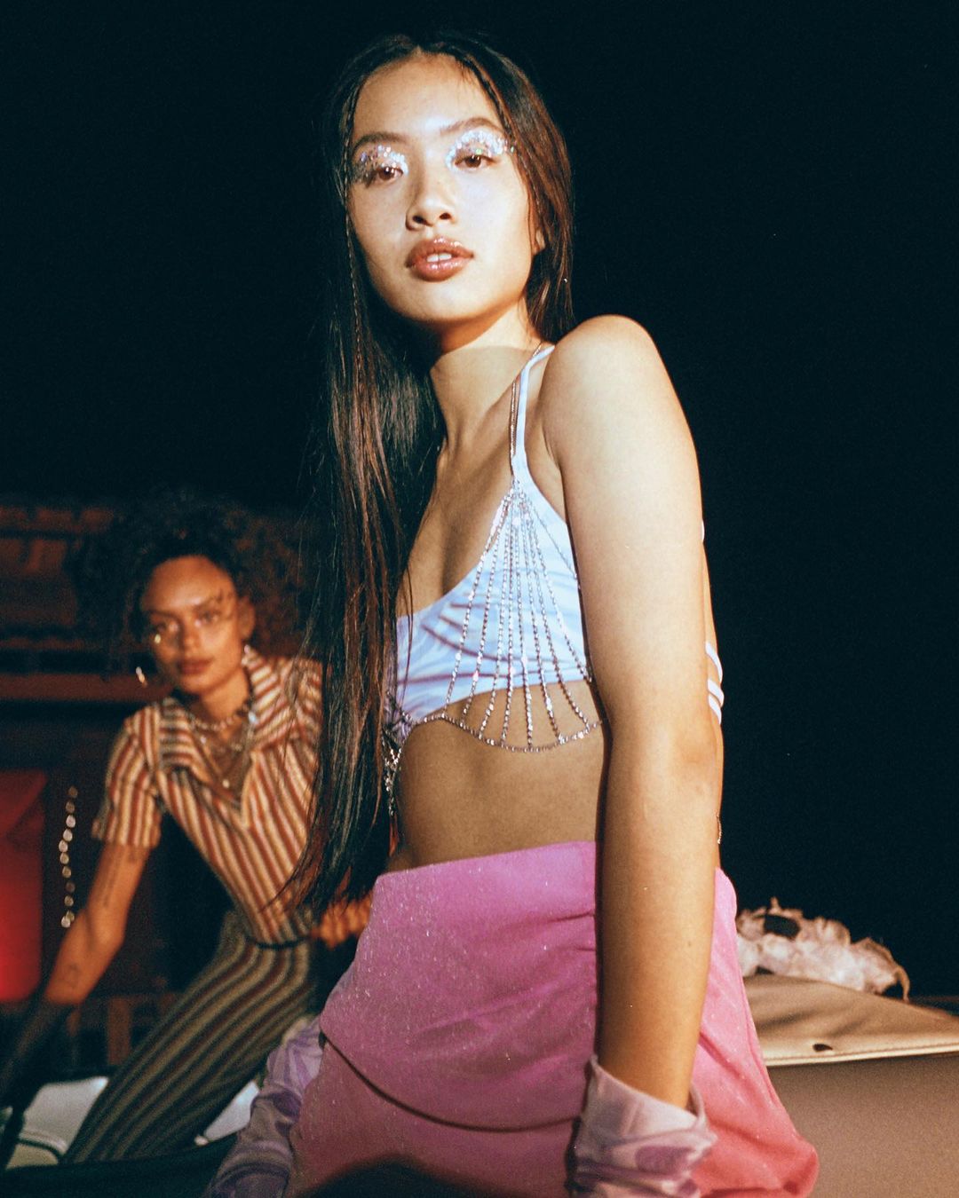Two young women pose indoors; one in a sparkly top and pink skirt in the foreground and the other in a striped top in the background.
