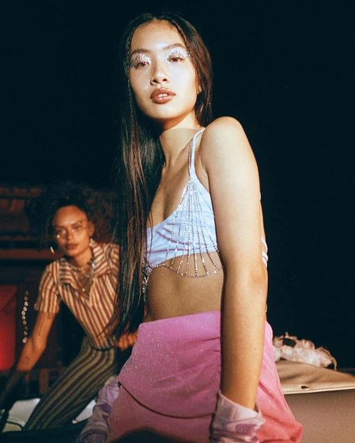 Two young women pose indoors; one in a sparkly top and pink skirt in the foreground and the other in a striped top in the background.