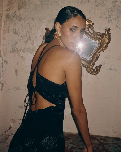A young adult female model with dark hair poses looking over her shoulder in a black dress in front of a gold framed mirror.