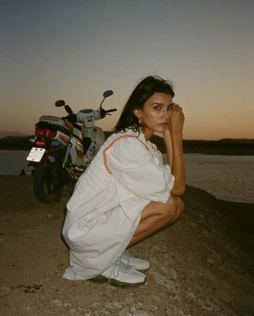Woman poses with motorcycle near body of water at sunset, wearing white dress and vest, sandy environment with neutral tones.