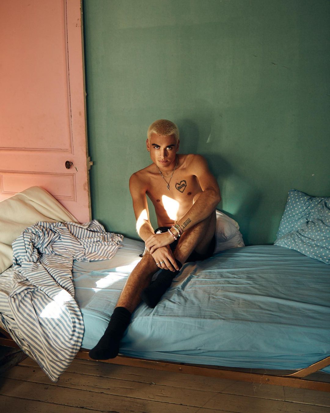 Young adult male poses shirtless on a blue bed in front of a pale green wall next to a pink door. The room is brightly lit.