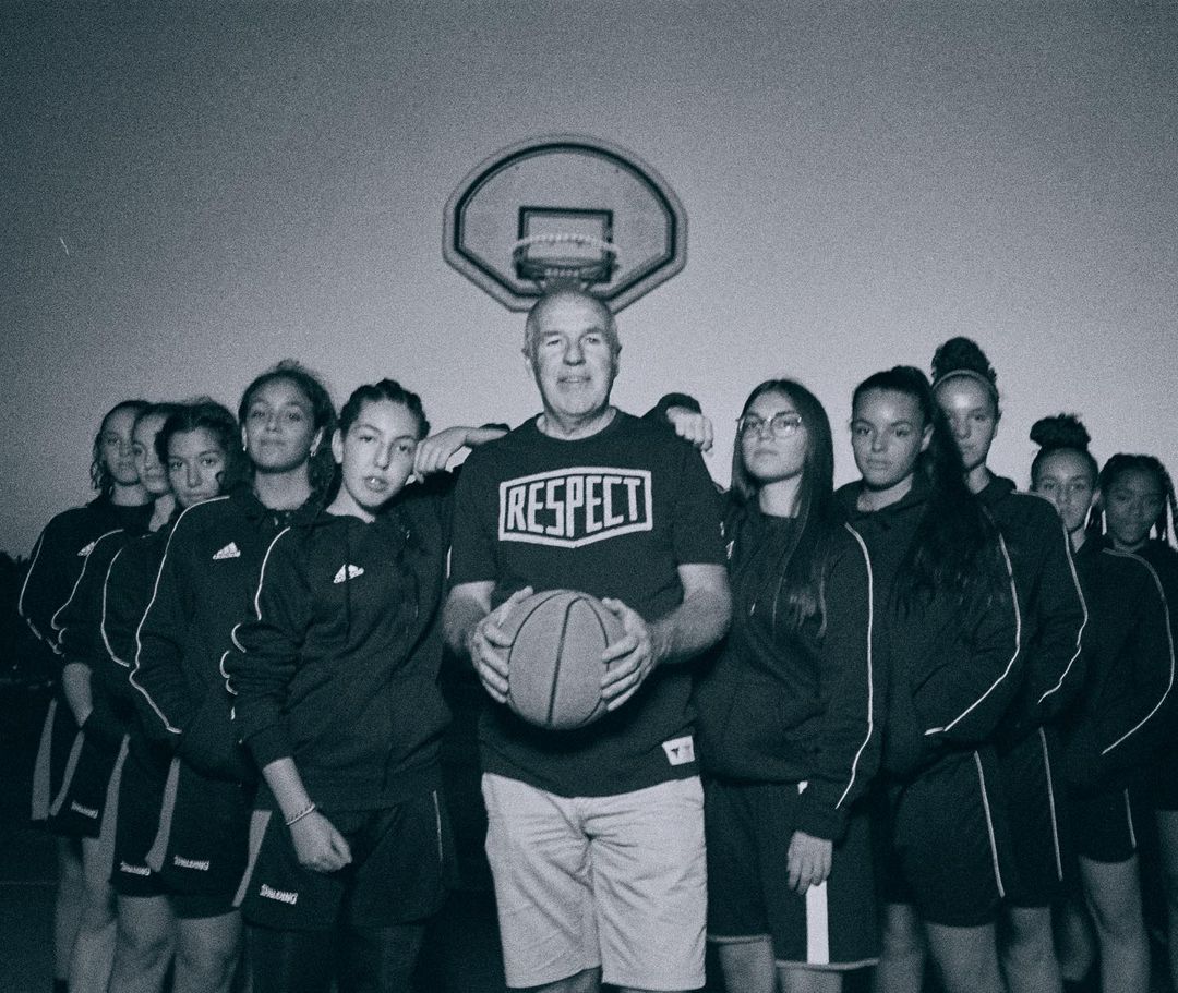 Team portrait of a girls' basketball team posing with their coach, wearing a RESPECT t-shirt, holding a basketball, in an indoor gymnasium.