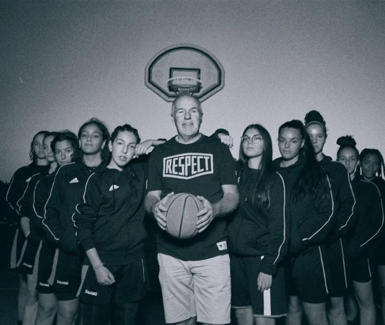 Team portrait of a girls' basketball team posing with their coach, wearing a RESPECT t-shirt, holding a basketball, in an indoor gymnasium.