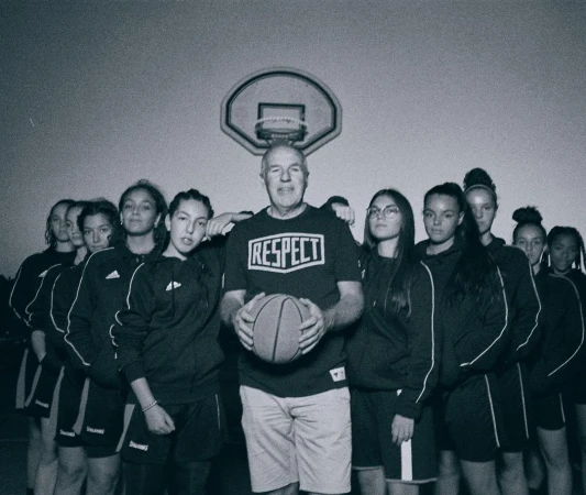 Team portrait of a girls' basketball team posing with their coach, wearing a RESPECT t-shirt, holding a basketball, in an indoor gymnasium.