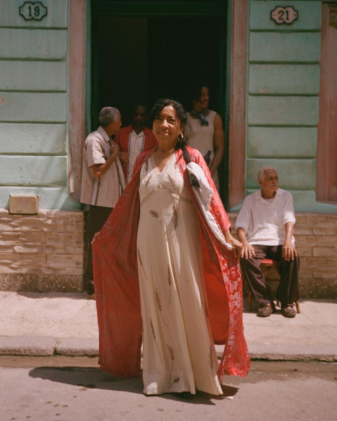 A woman in a long dress and robe stands smiling in front of a doorway, with other people and a building facade in the background.