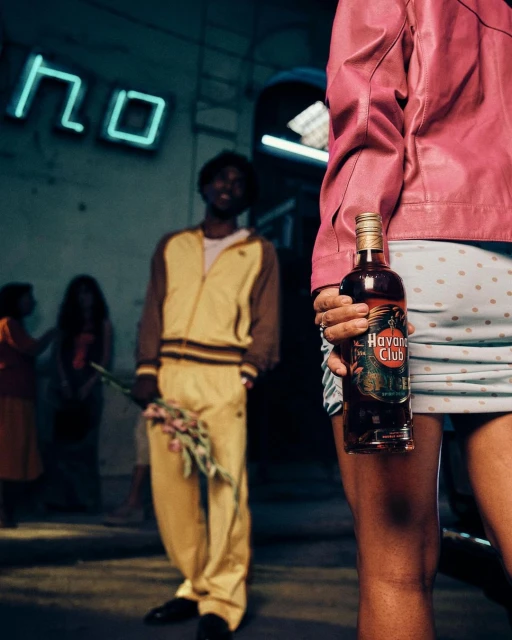 Woman holding a bottle of Havana Club rum, with a man holding flowers and a neon sign visible in the outdoor background. 