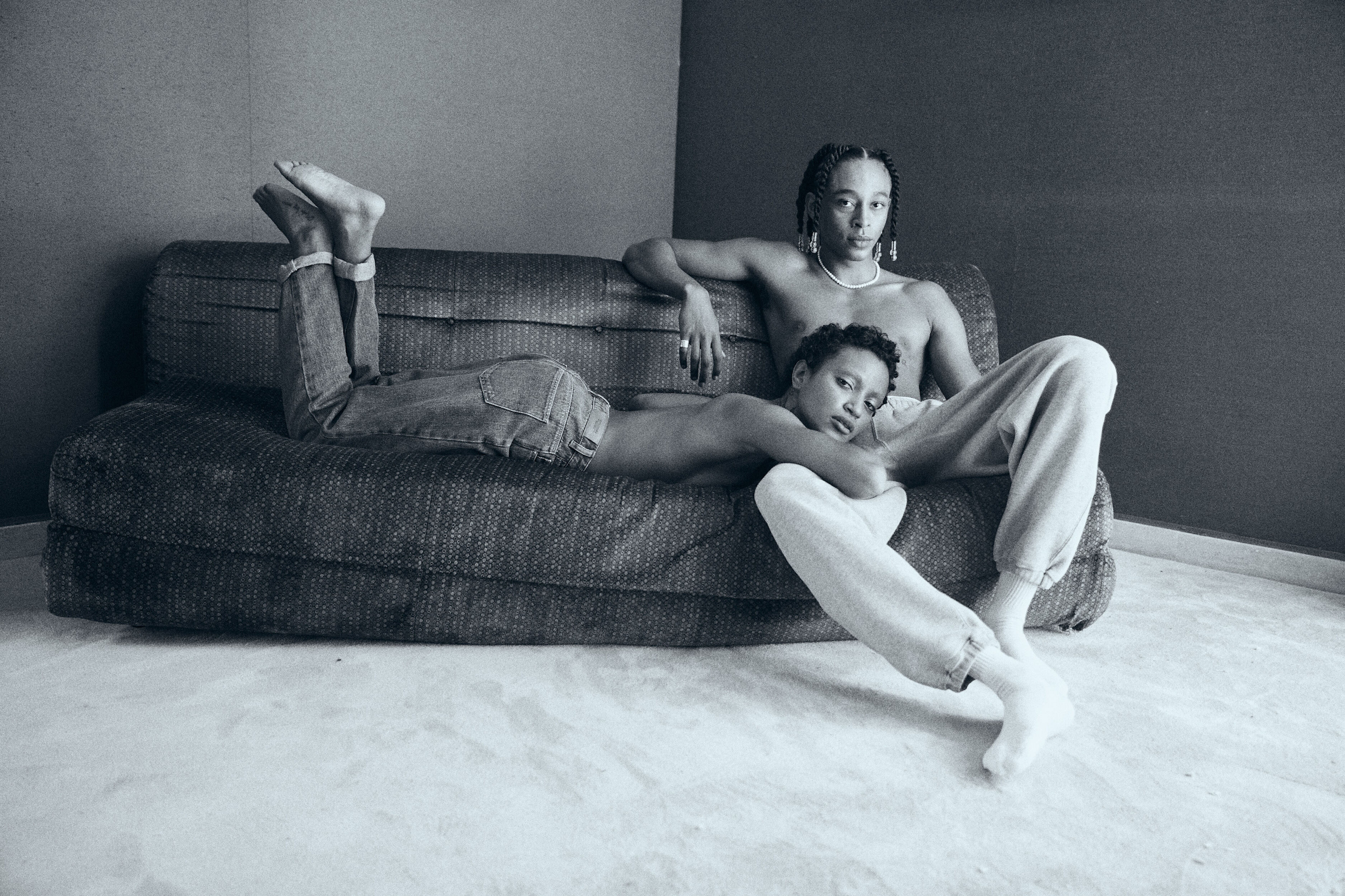 Black-and-white portrait of two shirtless people, one lying on a sofa and the other sitting, in an indoor setting.