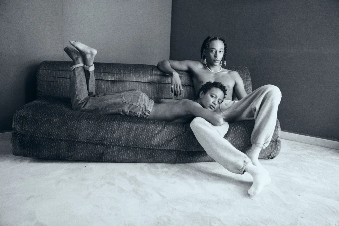 Black-and-white portrait of two shirtless people, one lying on a sofa and the other sitting, in an indoor setting.