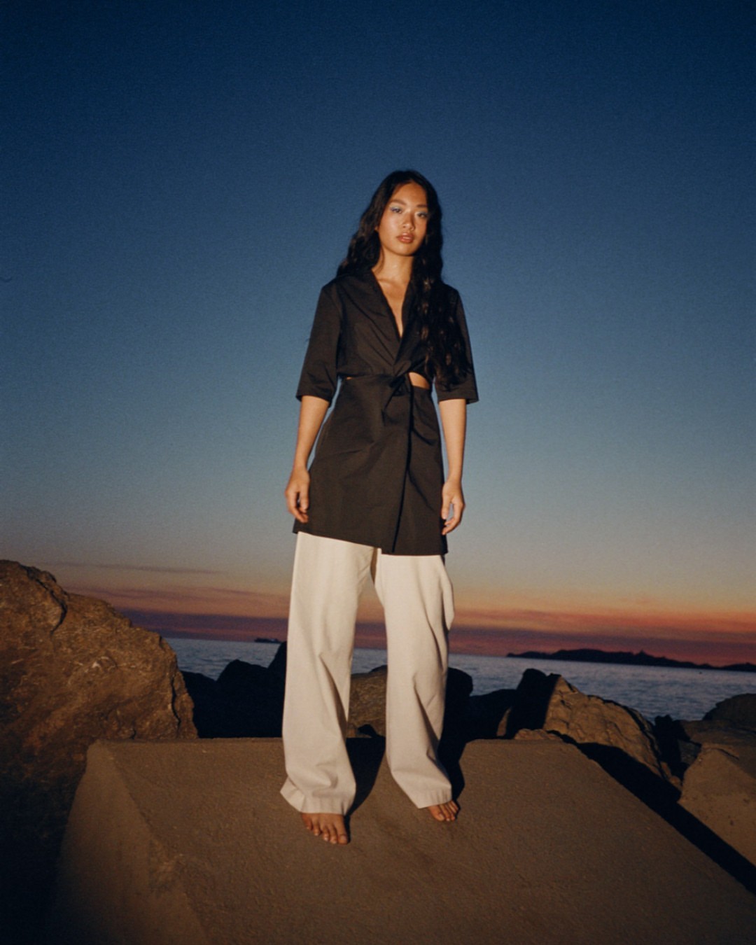 Woman in wide-leg pants and wrap top stands on rocks at dusk with a gradient sky as the sun sets over the ocean.