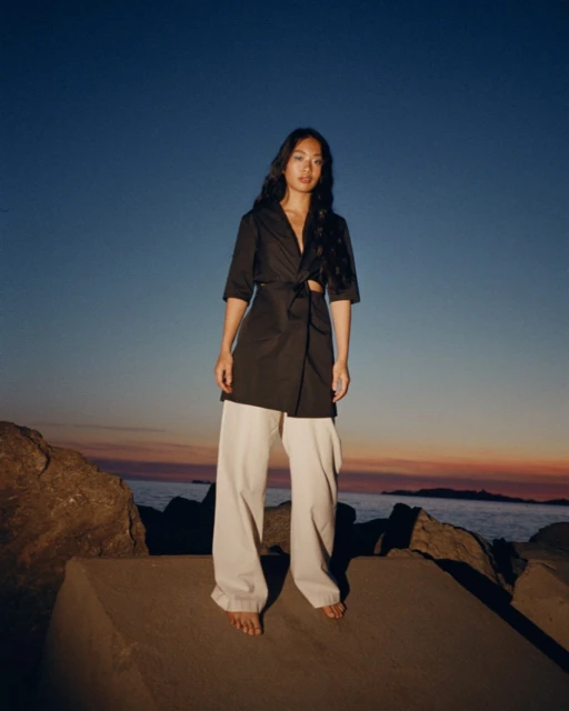 Woman in wide-leg pants and wrap top stands on rocks at dusk with a gradient sky as the sun sets over the ocean.