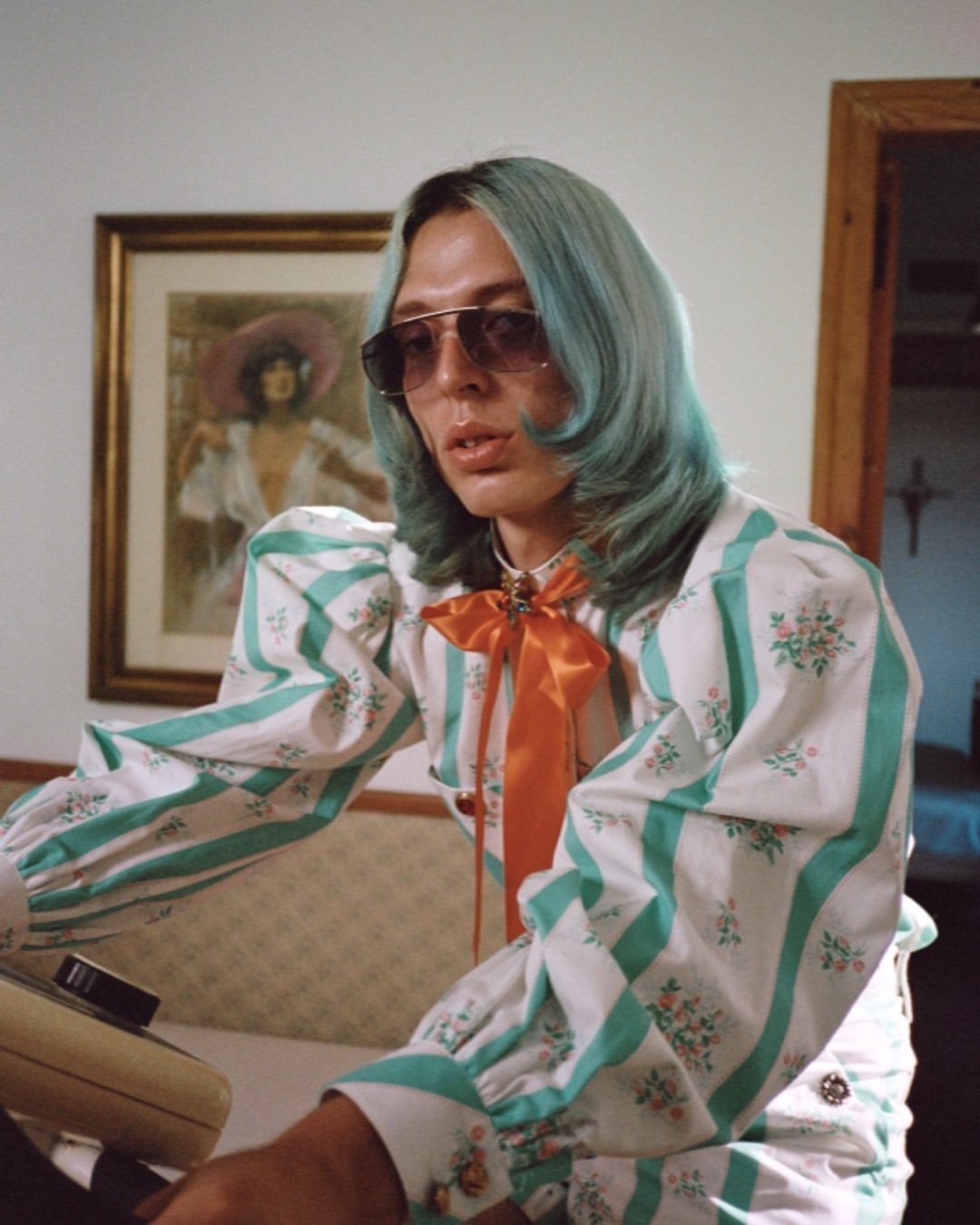 Person with blue hair, sunglasses, and floral striped dress poses indoors with artwork and vintage telephone in background.