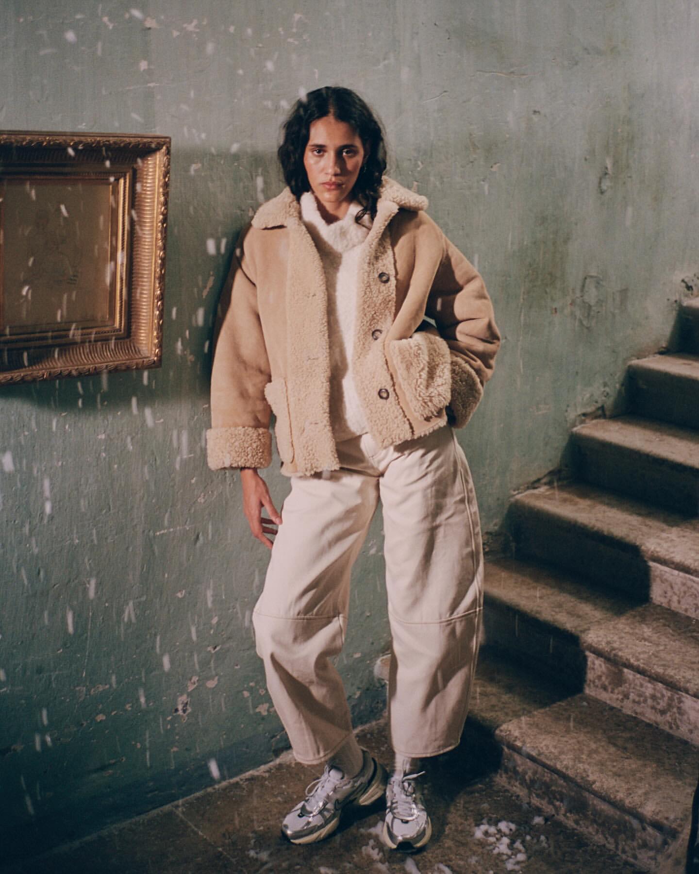 Woman poses indoors wearing tan shearling jacket, white sweater, pants, and running shoes, framed by a gold picture frame and stairs.