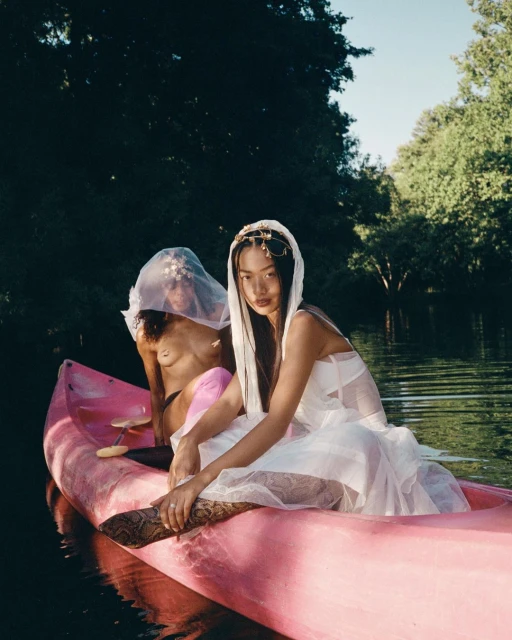 Two women in a pink canoe on a lake, wearing white dresses and veils, surrounded by green trees under a bright blue sky.