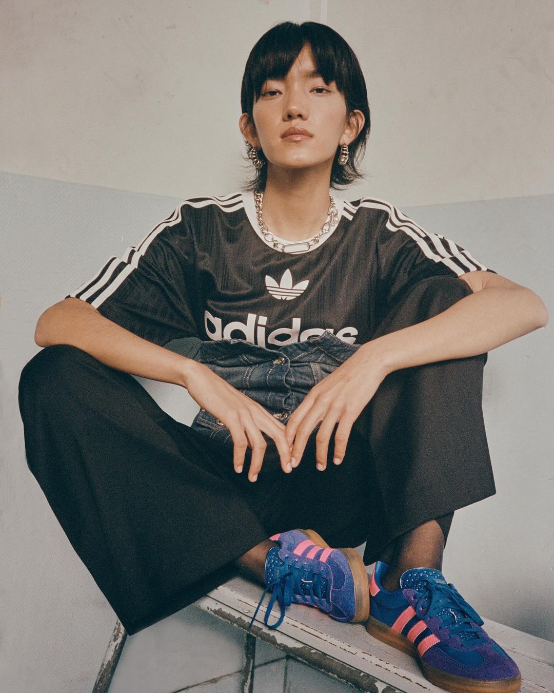 A woman in an Adidas shirt and wide-leg pants sits on a step stool wearing Adidas sneakers, posing in a bright indoor setting.