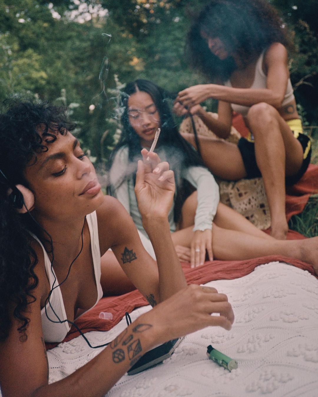 Three young women relaxing outdoors on a blanket. One lies down listening to music, while the other two sit up. 