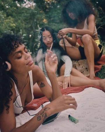 Three young women relaxing outdoors on a blanket. One lies down listening to music, while the other two sit up. 