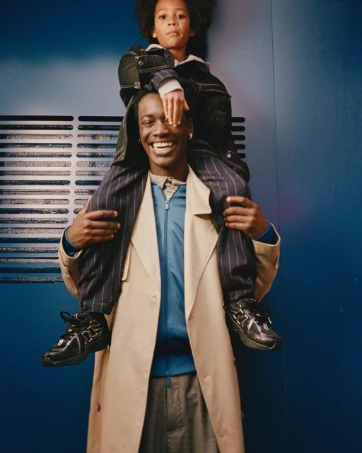 A smiling man carries a child on his shoulders in a studio shot. The child covers his own eyes. Both wear stylish jackets.