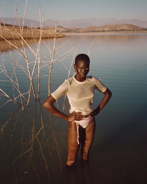 Elegant woman stands waist-deep in water, posing with hands on hips. Background features lake, distant mountains, and sparse trees.